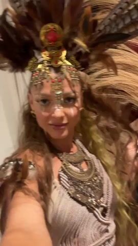 This media file is a close-up, selfie-style video of a woman dressed in an elaborate, tribal-inspired costume. She is wearing a large headdress adorned with feathers, gold metalwork, and red jewels. Her face is painted with white markings around her eyes and nose, and she has intricate gold jewelry on her forehead. She is also wearing a beaded, macrame-style top and a heavy, multi-layered gold necklace. Her hair is long and wavy, with some strands appearing to be dyed blonde.

The woman is smiling and looking directly at the camera, with her right hand held up and her fingers splayed, as if gesturing or posing. The background is softly lit and appears to be indoors, with hints of dried palm leaves and some white decorative vases visible. The overall aesthetic suggests a costume party, festival, or a themed photoshoot. The woman's expression is playful and confident. There is no visible text in the media file. The context provided indicates the location is Ko Samui District, Thailand, suggesting the setting might be a resort or event venue in this tropical location.