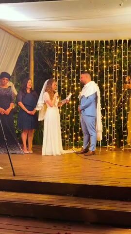 A wedding ceremony is underway under a white canopy adorned with bamboo poles. A bride in a white wedding dress and veil stands to the left, holding a piece of paper and speaking into a microphone. Facing her on the right is the groom in a light blue suit, also wearing a tallit (Jewish prayer shawl). Behind the bride stand two women, one in a dark blue dress and the other in a darker, embellished dress. Behind the groom, a person in a light-colored suit appears to be recording the event. The backdrop consists of a wall of hanging fairy lights. The scene takes place on a wooden platform.