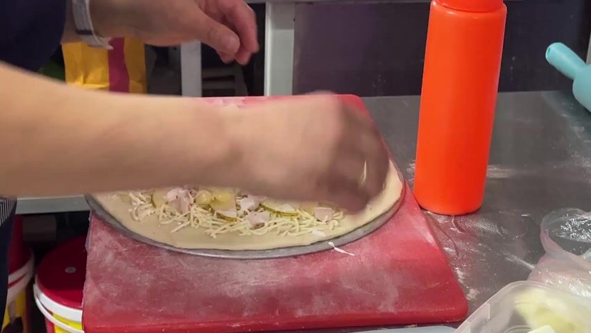 A person is making a pizza. The pizza has already been prepared with cheese, sliced pickles and chicken. The person is now adding cubed pieces of white cheese from a plastic container to the pizza. The pizza is on a metal pan, which is on top of a red cutting board. A red squeeze bottle, yellow bottle, and blue rolling pin sit near the pizza. The countertop appears to be metal. The person has a white watch on their left wrist and a ring on their right hand.