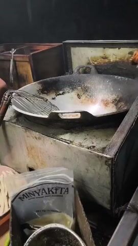A person is deep frying food in a wok. The wok is on a stove with a metal grate in front of it. The person is wearing a grey hooded sweatshirt and shorts. There is a bag of cooking oil on the bottom right of the frame. The background is dark.