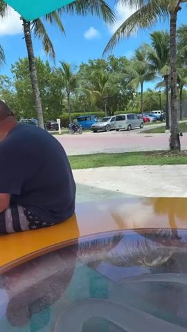 A man is sitting on a yellow surface, facing away from the camera, under a teal canopy supported by palm trees. He is wearing a dark blue t-shirt and American flag-patterned shorts. A black plastic bag filled with items sits to his left. In the background, there is a paved area with parked cars, including a blue van and a silver SUV, and lush green trees under a bright blue sky with scattered clouds.