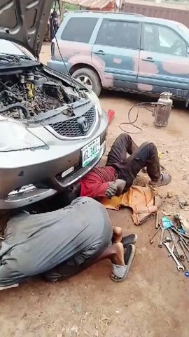 The video depicts two mechanics working on a silver Honda car in an outdoor setting in Karshi, Nigeria. The car's hood is open, indicating engine or front-end work.

In the initial frames (0:00-0:02), both mechanics are lying on their backs on the ground, positioned under the front of the car. One is directly under the center, while the other is closer to the front left wheel, appearing to be focused on the underside of the vehicle. Various tools are scattered on the dirt ground around them. A gas cylinder with a hose is visible in the background, near a blue SUV (likely a Honda CRV) which is also parked in the open area. The silver car displays a Nigerian license plate that reads "LAGOS AGL-341CC".

As the video progresses (0:02-0:05), the camera pans slightly to show the front left side of the car. One mechanic is now kneeling beside the front left wheel, actively working within the wheel well area. The other mechanic remains lying on his back on the ground, in the foreground. Near him, an oil pan containing dark liquid (presumably used engine oil) and a brake drum are visible, suggesting brake or suspension work in addition to potential engine issues.

The setting appears to be an informal, open-air auto repair shop or a roadside repair location. The ground is dusty dirt, and the scene is illuminated by daylight. The mechanics are focused on their tasks, demonstrating a typical daytime vehicle repair operation.
