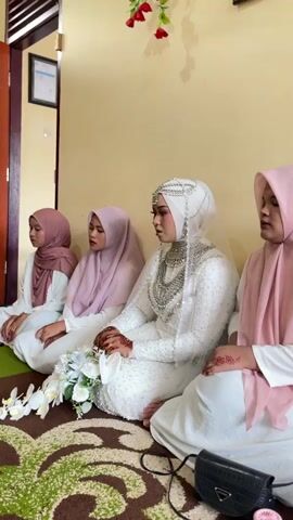 A group of four women are sitting on the floor, facing the camera. The woman in the center is wearing a white wedding dress and hijab, with elaborate silver jewelry around her neck and on her head. She is holding a bouquet of white flowers. To her left are two younger women, also wearing hijabs. To her right is an older woman wearing a pink hijab. All women are looking down or to the side with solemn expressions. The room appears to be a home, with a yellow wall and a wooden door in the background. A patterned rug is on the floor. A black purse and a pink object are on the floor in front of the women. The scene seems to be part of a religious ceremony or a pre-wedding ritual.