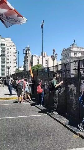 The video shows a protest or demonstration taking place in an urban setting. Initially, a metal barricade is visible, with people standing near it. The camera then moves to show a crowd of people marching down a street. Many are carrying flags and banners, including some with red and white colors and others with light blue and white, resembling the Argentinian flag. The banners feature text that appears to be in Spanish. Some people are holding long poles, and the general atmosphere seems energetic. Buildings line the street, and the sky is clear and blue. The crowd is diverse in age and appearance, and they are all moving in the same direction. The overall scene suggests a political rally or protest.