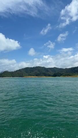 The video shows a body of water, possibly a lake or reservoir, with rolling hills covered in dense green foliage in the background. The sky is blue with scattered white clouds. The water is a deep turquoise color and appears to be slightly choppy. The camera is positioned low, close to the water's surface, suggesting it might be mounted on a boat or other watercraft. There are no people or other obvious objects visible in the frame. The overall impression is one of a natural, serene landscape.