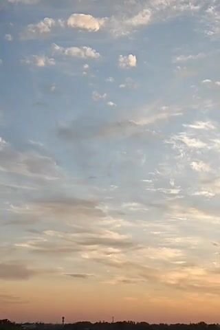 A time-lapse video shows clouds drifting across a pale blue and orange sky at sunset. The lower portion of the frame shows a dark silhouette of trees and a few structures against the horizon.