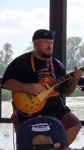 The video is a medium shot of a bald, white man playing an electric guitar. He is wearing a black beanie, sunglasses perched on top of it, a black t-shirt, and maroon shorts. He is seated, and the guitar is an orange and yellow sunburst Gibson Les Paul. He is actively playing the guitar, strumming and fretting. The background shows a body of water and trees, suggesting an outdoor or semi-outdoor setting. In the foreground, a blue baseball cap with a white logo is partially visible. The lighting is natural, indicating daytime.