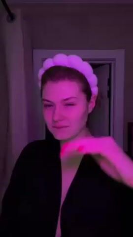 A young woman in a black robe is in a bathroom with pink lighting. She has reddish-brown hair and a light complexion. She is wearing a fluffy, white headband. She adjusts the headband, removes it, and then runs her hand through her hair, pushing it behind her ear. A white shower curtain and a partially open white door are visible in the background.