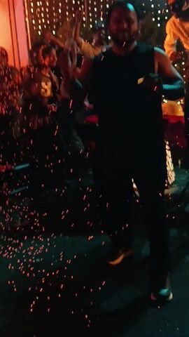 The video shows a group of people at an outdoor event, possibly a party or celebration. The scene is filled with a shower of sparks and a burst of flames from a pyrotechnic effect. A man in a black tank top and pants is prominently featured in the foreground, seemingly enjoying the spectacle. Other individuals are visible in the background, some dancing and others holding objects like a trumpet. The environment is illuminated with decorative string lights. The ground is littered with sparks from the pyrotechnics, creating a visually dynamic and festive atmosphere.