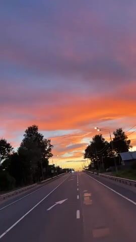 The video captures a road scene at sunset in Kuzhenkino, Russia. The sky is ablaze with vibrant orange and pink hues, a stunning display of a sunset. The road is asphalt, divided by a white dashed line and a solid white line on the right. A white arrow is painted on the left lane, indicating a turn.

On either side of the road, there are trees and streetlights. A few cars and trucks are visible on the road, moving away from the camera. The atmosphere is serene, with the setting sun casting a warm glow. The time of day is clearly sunset, as indicated by the dramatic colors in the sky. The location cues are the road and the trees, typical of a rural or suburban setting. The overall mood is peaceful and picturesque, enhanced by the beautiful sunset. There are traffic signs visible, but no discernible text can be read.