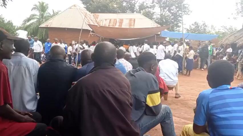 The video shows a crowd of people gathered outside a building. In the foreground are seated individuals watching the scene unfold. In the middle ground, a larger group of people, mostly children dressed in school uniforms, are standing. The children are arranged in a loose formation and are looking in different directions. Behind them, there's a small building with a corrugated metal roof, shaded by a large cloth canopy. The environment appears to be a rural area, with trees and foliage visible in the background. The video seems to capture a social gathering or event, possibly related to the school children.