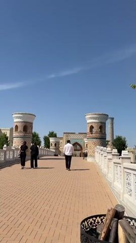 The video shows three people walking away from the camera along a wide, paved pathway on a bright, sunny day. The pathway, made of reddish-brown interlocking pavers, leads towards an ornate arched gateway flanked by two large, circular towers. The towers and the gateway feature intricate brickwork and decorative blue and white tile patterns. White, decorative railings line both sides of the pathway. Beyond the gateway, more architectural elements and some small trees are visible. The sky above is clear and blue with minimal clouds. In the foreground, a dark, decorative metal object, possibly a fire pit or planter, is partially visible on the right. The scene depicts an outdoor architectural space, likely an entrance or bridge, in Kuchtumgali, Uzbekistan.