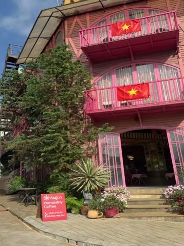 The video captures various street scenes in Măng Đen, Vietnam, featuring charming, architecturally distinct buildings and a clear, sunny day.

The initial shots showcase a paved street flanked by multi-story buildings. These structures often feature dark wooden facades with prominent balconies, some painted green and others red. Many red balconies are adorned with festive red ribbons or streamers. Large potted plants filled with white and purple flowers line the street, enhancing the aesthetic. The sky is bright blue with scattered white clouds, indicating clear daytime conditions.

In the first shot, two individuals, one clearly visible in black attire, walk down the street past a golf cart-like vehicle. A white SUV is parked further ahead. The second and third shots provide wider views of the street and buildings, showing a repetitive pattern of the rustic architecture. A brown SUV and a black pickup truck are also visible among parked vehicles.

A close-up focuses on the entrance of one building, identified by the text "Cozy House" above its doorway. A Vietnamese flag (red with a yellow star) hangs above the entrance. Steps lead up to the building, with flower pots on either side, and a colorful mural is painted on an adjacent wall.

The final segment shows a narrower street lined with buildings of varied styles; one on the left has green balconies, while a white building on the right features pink balconies. A golf cart-like vehicle is in the foreground, and a white Hyundai Santa Fe SUV with license plate "30K-601.51" is parked. Overhead power lines are visible, completing the streetscape. The overall atmosphere suggests a pleasant, possibly tourist-oriented, village or resort area.