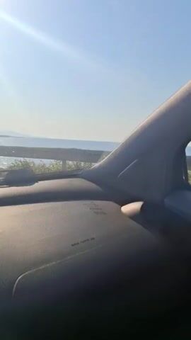 The video shows a view from inside a vehicle driving along a coastal road. To the right of the road is the sea, glittering under a bright sun. The sun is high in the clear blue sky. A metal guardrail separates the road from the coastline. In the distance, land is visible. The dashboard of the vehicle occupies the lower portion of the frame.
