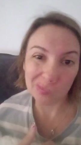 A woman with light brown hair, wearing a striped shirt, is facing the camera. She purses her lips in a playful way, moving her head slightly and moving her index finger. She has brown eyes and appears to be sitting on a couch. The background is a plain white wall.