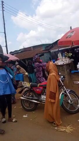 A short video captures a bustling street market scene in what appears to be a developing country. The footage begins with a child carrying a tray on their head, followed by a woman. The camera pans to reveal numerous market stalls set up under umbrellas, displaying various goods. People, mostly women dressed in traditional attire including hijabs, are seen interacting with each other and attending to their stalls. Some stalls have buckets or containers filled with goods, possibly food items or produce. The ground is dirt and scattered with debris. Further into the video, a woman is seen washing clothes in a large black basin. Motorcycles are parked haphazardly, and a man sits on a cart laden with corn. A large political banner with the text "RENEWED HOPE AGENDA 2027" and images of several men is prominently displayed. The overall impression is of a vibrant, albeit somewhat disorganized, local marketplace.
