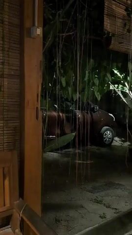 The video shows an outdoor seating area at night. A brown car is parked in the background behind some hanging vines. In the foreground, there are wooden benches and tables. The lighting is dim, with some green lights visible in the upper right corner. The environment appears to be a tropical or garden setting, with lush greenery behind the car.