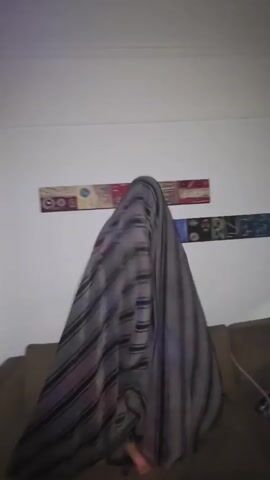 The video shows a person under a large striped blanket in a living room. The person, wearing red plaid pants, is standing near a brown sofa. Behind them is a white wall with a framed picture. The music plays loudly.