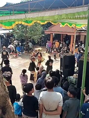 This video shows an outdoor gathering where a traditional performance is taking place. A group of people dressed in colorful costumes are engaged in a dance or ritual in the center of an open area. Some are wearing masks and elaborate headdresses, with one performer holding a long stick. Spectators are gathered around, watching the performance. In the background, there are buildings, trees, and a covered structure decorated with green and yellow banners. There are also sound equipment, including speakers, set up to the right of the performance area. The ground appears to be paved and wet in some areas, suggesting recent rain or water. The overall atmosphere seems festive and communal.