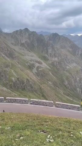 The video captures a scenic mountain landscape with a winding road. The road has a stone retaining wall with a black and yellow striped pole on the right side of the frame. The road curves to the right and ascends into the mountains, where more roads and some structures, possibly buildings, can be seen in the distance. The mountains are covered in green vegetation and rocky terrain, with a cloudy sky overhead. There are no people clearly visible in the frame.