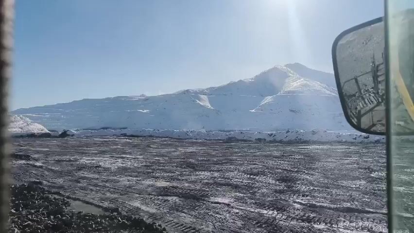 The video shows a snow-covered landscape from the inside of a vehicle, possibly a truck or heavy machinery, given the view from the cab. The foreground is a field or open area with tire tracks in the dirt or snow. In the background, there is a large, snow-covered mountain with visible levels or terraces, suggesting it might be a quarry or mining operation. The sky is bright and blue, and the sun is shining. A side mirror of the vehicle is visible in the frame.