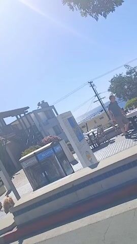 The video shows a street scene on a sunny day. There is a building with a sign indicating a bus stop. The sign has the number 438 and 109. There are people walking around, including a child in a blue shirt and red pants. In the background, there is a view of the ocean. There are trash and recycling bins as well.