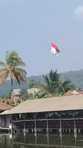 A red and white flag flutters on a flagpole against a clear sky. In the foreground, a wooden structure with a brown roof and supporting posts is partially submerged in water. Trees, including a palm tree, are visible behind the structure. In the background, rolling hills covered in greenery are seen. The flag appears to be the Indonesian flag. The overall scene depicts an outdoor environment, possibly a rural or semi-rural area.