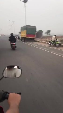 The video shows a first-person perspective of someone riding a motorcycle on a road. The rider's hand is visible on the handlebar and the side mirror reflects the rider's upper body wearing a white shirt. In the distance, other vehicles including motorcycles, cars, and a truck with a green tarp are on the road. The environment appears to be somewhat hazy, possibly due to weather conditions, and there are streetlights lining the road. The road itself is paved, with a shoulder and some construction or undeveloped land to the right. The vehicles appear to be moving in the same direction as the motorcycle.