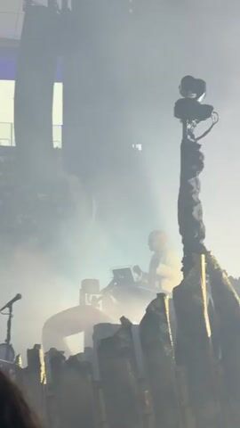 The video shows a person in a white jacket and glasses at a keyboard on a stage. There is fog and stage lights creating a dramatic atmosphere. A tall, dark structure with a camera on top is to the right of the person. The background is blurry and dark with some lights visible.