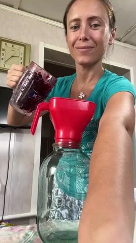 A woman with dark hair pulled back is pouring a dark purple liquid with solid pieces from a glass pitcher into a large glass jug through a red plastic funnel. She is wearing a teal t-shirt and a patterned skirt. The background shows a wall with a clock and a doorway.