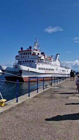 A white ferry named ONYX is docked at a pier on a sunny day. The ferry has multiple decks and is painted with red accents along its waterline and on life preserver rings visible on the side. The name ZEG LUG A GDAŃSKA is written on the side of the ferry. A blue railing runs along the edge of the pier. A person is walking away from the camera on the pier. The sky is clear blue with a few wispy clouds.