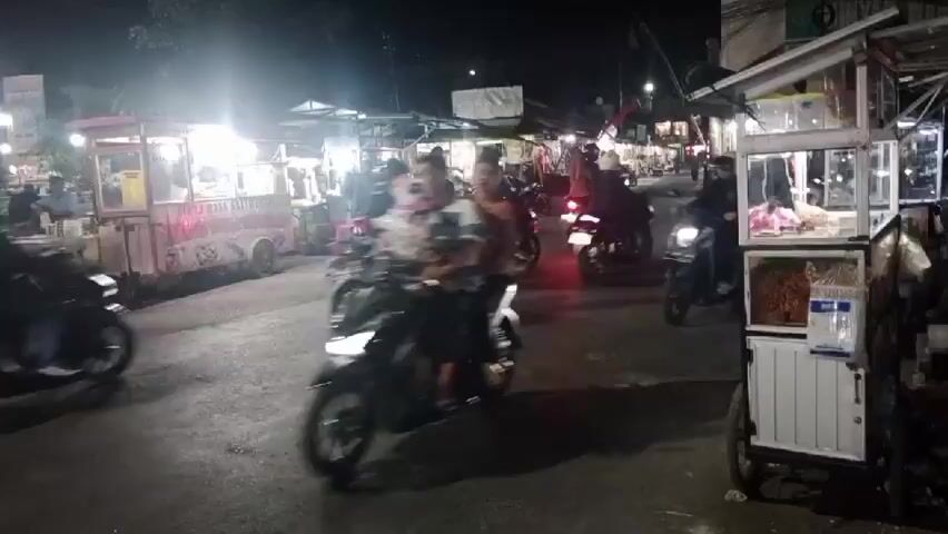 This is a nighttime video of a busy street market. Motorbikes are the primary mode of transportation, with many of them driving through the market, creating a dynamic and somewhat chaotic scene. Several food stalls are visible along the sides of the street, illuminated by artificial lights. People are seen operating and patronizing these stalls. The overall environment is dark, with the lights from the stalls and motorbikes providing the main illumination. The camera appears to be mounted on a moving vehicle, likely a motorbike, as evidenced by the slight blur and the perspective of the footage.