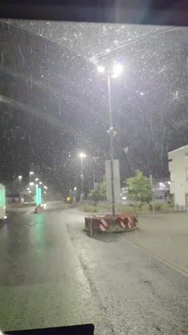 This is a nighttime, exterior shot looking through a windshield that is wet with rain. The rain is visible as streaks and droplets hitting the glass. The scene shows a paved road with streetlights illuminating the wet asphalt and surrounding area. There are some green illuminated bollards on the left side of the road, a traffic island with red and white barriers in the middle of the road, and a building on the right side. Trees are also visible in the background. The overall impression is of a rainy night scene, likely in an urban or semi-urban environment.