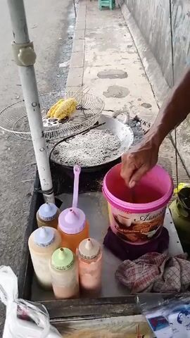 A man is preparing street food on a cart next to a sidewalk. He is stirring something in a pink bucket with a spoon and cooking strands of something in a large pan of hot oil. A corn on the cob sits in a wire basket to the side of the pan. A tray on the cart holds multiple bottles of sauces and condiments. A green stool sits on the sidewalk in the background.