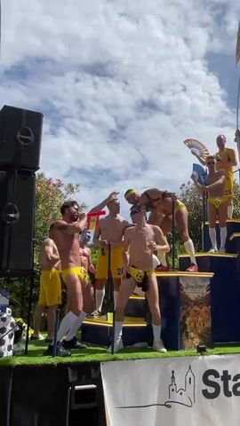 The video shows a group of men on a float decorated with a Spanish flag and a large trophy. They are all wearing yellow swimsuits and white socks. The men are dancing and cheering, and one of them is holding a flag with the words "Stay Sitges" on it. The float is decorated with images of soccer players and a rainbow flag. The sky is blue with white clouds.