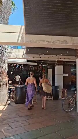 The video shows a courtyard area with an outdoor seating section of a building, possibly a bar or restaurant. Two women walk away from the camera towards the building. One woman is wearing a long purple dress, and the other is wearing a dark dress and carrying a large straw bag. They are walking on a paved area with a palm tree on the left side of the frame. The building has an awning over the seating area, decorated with string lights. Inside the building, there are tables and chairs, and several people are visible. The atmosphere appears to be daytime and sunny.