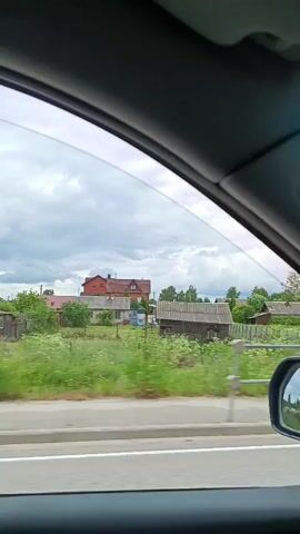 The video captures a view from inside a moving car, looking out the passenger-side window. The scene outside is a rural landscape in Savinskaya, Russia, under a cloudy sky. The time of day appears to be daytime, given the ambient light.

The view through the window shows a series of houses and other structures, characteristic of a village or countryside. There are several buildings with pitched roofs, some appearing to be wooden sheds or outbuildings. Green vegetation is abundant, with grassy fields and trees filling the middle ground. A road with a white line marking is visible in the foreground, separated from the fields by a metal guardrail. The car's side mirror is also visible on the right side of the frame, reflecting the passing scenery and sky.

The overall atmosphere is that of a journey through a quiet, perhaps remote, Russian countryside. The cloudy sky suggests possibly overcast or showery weather, but it's not overtly raining. There is no visible text in the media. The focus is on the passing scenery and the experience of traveling through this environment.