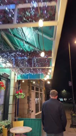 A man walks towards the street at night. The street is lit by streetlights and the lights of a building that houses “Temple Heelis Solicitors”. The building has a green awning above the entrance, decorated with small lights and artificial foliage.  A round table and a wooden chair are in the foreground next to a green privacy screen. There is a flower basket hanging from a post.