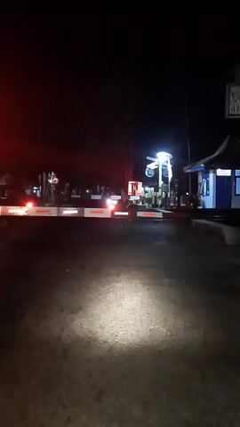 The video shows a dark night scene at a railroad crossing. The gate is down, blocking traffic, indicated by red flashing lights and white and red striped barriers. A bright spotlight from a vehicle illuminates the road directly in front of the camera. In the background, a small building with blue siding and a dark roof is visible, possibly a ticket booth or station office. A white pole with lights and what appears to be a railway signal mechanism is also present. The overall atmosphere is one of waiting, likely for a train to pass.