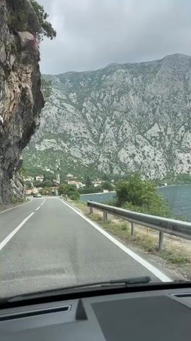 The video captures a first-person perspective from inside a moving vehicle, showcasing a scenic drive along a coastal road in Donji Orahovac, Montenegro. The view is dominated by a dramatic landscape featuring a vast, rugged grey mountain range on the left and spanning the background, covered intermittently with sparse green vegetation. To the right of the asphalt road, a calm, dark blue-green body of water, likely a bay or lake, stretches out.

The road itself is a two-lane asphalt surface with clear white markings, curving gently to the right. A metal guardrail separates the road from the water's edge on the right. On the left, the mountain rises sharply, with a section of rocky cliff face almost overhanging the road.

In the mid-ground, nestled at the base of the mountains along the water, is a small cluster of buildings with distinct red roofs, characteristic of coastal settlements in the region. A tall, grey cell tower or antenna structure stands prominently among these buildings. Another white car is visible driving in the distance on the same road.

The sky overhead is overcast, indicating a cloudy daytime setting, possibly late morning or afternoon. The overall impression is one of a picturesque and tranquil journey through a beautiful natural environment. No specific people or visible text are discernible in the footage.