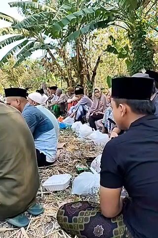 A group of people are sitting on the ground outdoors, appearing to be at an informal gathering. They are mostly dressed in traditional Muslim attire, including peci (caps). Some are facing away from the camera, while others are looking towards the center of the group. There are several white Styrofoam food containers and plastic bags scattered on the ground in front of them, suggesting a meal is being shared or has recently been consumed. The setting appears to be in a rural area, with trees and foliage visible in the background. The ground is covered with dry grass and straw. The lighting suggests it is daytime.