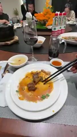 Here is a description of the video:

The video shows a table at a Chinese restaurant. There are several people sitting around the table, eating and talking. The camera focuses on a white plate with leftover meat and sauce. Black chopsticks are picking at the food. There is a glass of red wine next to the plate, along with other dishes and drinks. A decorative floral arrangement sits in the center of the table.