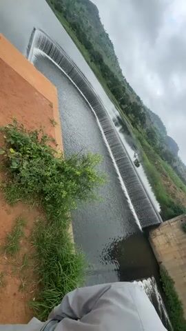 This video showcases a dam with water cascading over it. The dam is constructed of concrete and appears to be a curved structure. The surrounding environment is hilly and covered in greenery, with trees and vegetation on the slopes. The sky is overcast with clouds. There are no people visible in the video. The camera is positioned at an angle, giving a wide view of the dam and its surroundings. The water flow is continuous, creating a curtain of white water as it falls. The overall impression is one of a natural landscape with man-made infrastructure.