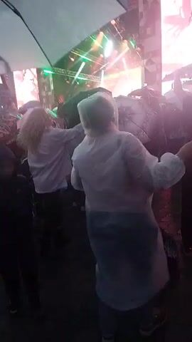 A person in a white hooded poncho is seen from behind, dancing at an outdoor music festival or concert. The person is facing a large stage with bright lights and screens. Other concertgoers are visible around the person, some holding umbrellas, suggesting it is raining or has recently rained. The overall atmosphere appears festive despite the weather.