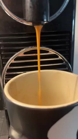 The image shows a close-up of an espresso machine dispensing coffee into a black and cream mug. The coffee, a rich amber color, flows in a thin, steady stream from the machine's spout, creating a slight ripple on the surface of the liquid already in the mug. The mug itself appears to be ceramic and has some light staining on the inside, suggesting it has been used recently. Behind the mug, the espresso machine is visible, primarily in black with a silver grille. The machine has a ribbed black surface above the grille. The background is a blurred kitchen counter. The overall impression is one of a domestic scene, possibly in the morning, given the activity of making coffee. There is no visible text in the image.