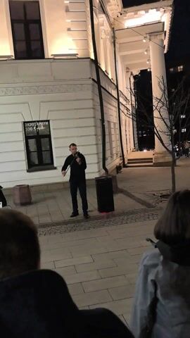 Here is a description of the video:

The video shows a man playing the violin in front of a building at night. The building is white with large columns and windows. The man is wearing a black shirt and pants. A black speaker stands next to him. People are standing in front of him watching and recording the performance.