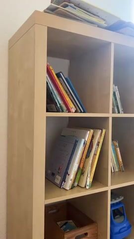 This video provides a panning view of a children's waiting room.  The room features a light-colored bookshelf filled with children's books, a blue toy truck, and a wooden crate on the bottom shelf.  To the right, a white bench with storage compartments is situated beneath a window. On the bench are several children's toys, including a colorful bead maze. The walls are painted teal and decorated with framed pictures of babies, posters, and blue wave-like accents. A row of blue chairs for seating is visible against the teal wall. A white radiator runs along the base of the window wall.