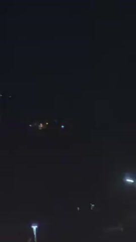 The video shows a dark night scene with distant lights that could be from a city or other development. The lights are blurred and indistinct, suggesting they are far away. Streetlights are visible, casting a glow in the immediate vicinity. There is a faint voice in the video.
