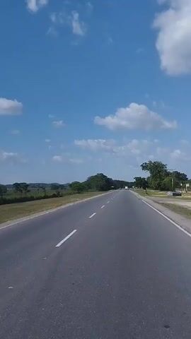 This video captures a view from a moving vehicle, likely a car, traveling along a paved road in Taguanes, Venezuela. The scene is dominated by a bright blue sky filled with fluffy white cumulus clouds, indicating a pleasant, sunny day. The road stretches ahead, marked with white lines, suggesting a highway or a major road.

To the left of the road, a grassy field extends, bordered by a dark fence. Beyond the field, a line of trees and distant hills form the horizon. The landscape appears rural and open, with vast stretches of green fields and scattered trees. The time of day is clearly daytime, likely late morning or early afternoon, given the bright sunlight and the clear blue sky.

There are no people or specific objects clearly visible in the foreground or on the roadside, except for the road itself and the natural elements. The activity taking place is simply travel along this road, with the camera providing a continuous, slightly shaky perspective of the passing scenery. The overall mood is peaceful and expansive, evoking a sense of journey and open space. No visible text is present in the media file.