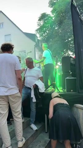The video captures an outdoor live music performance with a band on a stage. Several people are visible in the foreground, some facing the stage and others engaged in their own activities. One man in a white t-shirt and jeans is looking down, holding a white shirt. To his left, another man in a white t-shirt and light-colored pants stands facing away from the camera. In the background, a woman in a black dress and a light-colored hat bends down, holding a red bag. The band members are on stage, with a guitarist in a white t-shirt playing a guitar. Green and blue stage lights illuminate the scene, creating a smoky atmosphere. Trees and buildings form the backdrop.